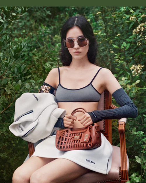 Woman sitting outdoors holding a brown handbag, wearing sunglasses and a gray top.